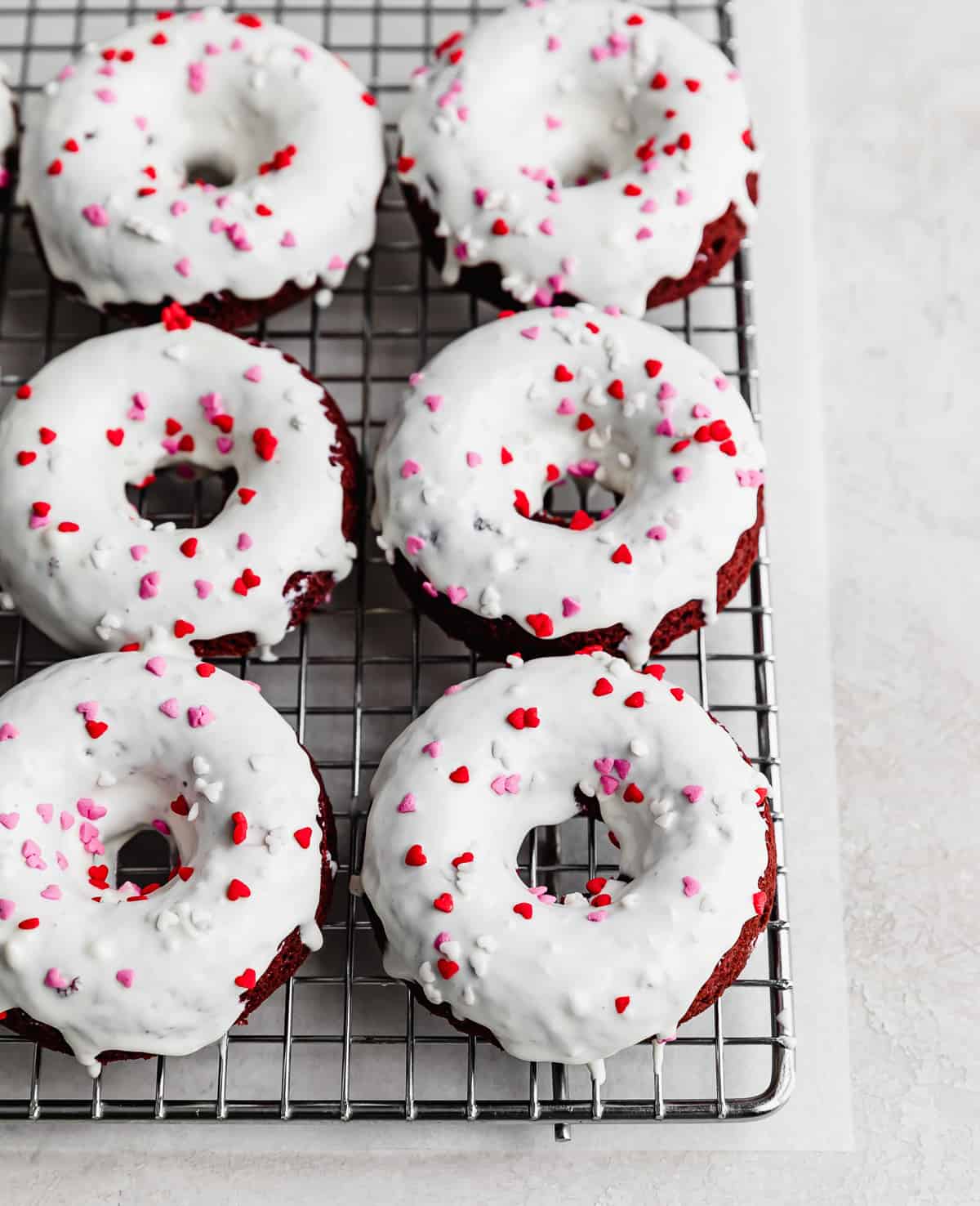 Baked Red Velvet Donuts topped with a white chocolate glaze and heart sprinkles on a wire cooling rack.