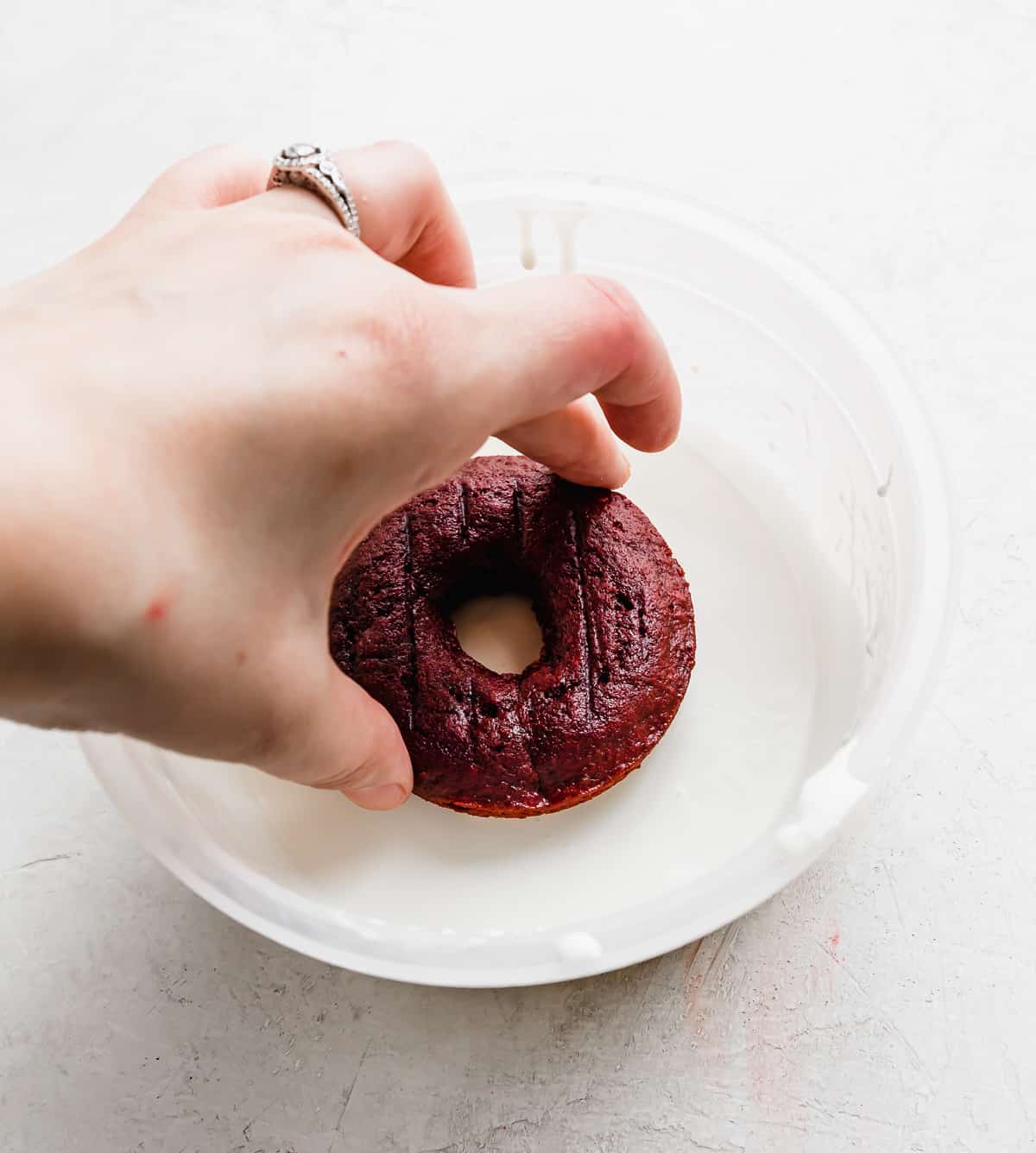A hand dipping a red velvet donut in a white chocolate glaze.