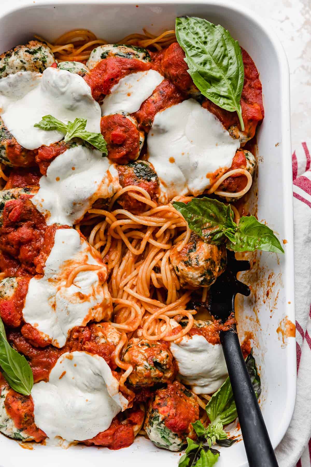 A white baking dish with marinara coated spaghetti noodles topped with turkey spinach meatballs and melted mozzarella cheese.