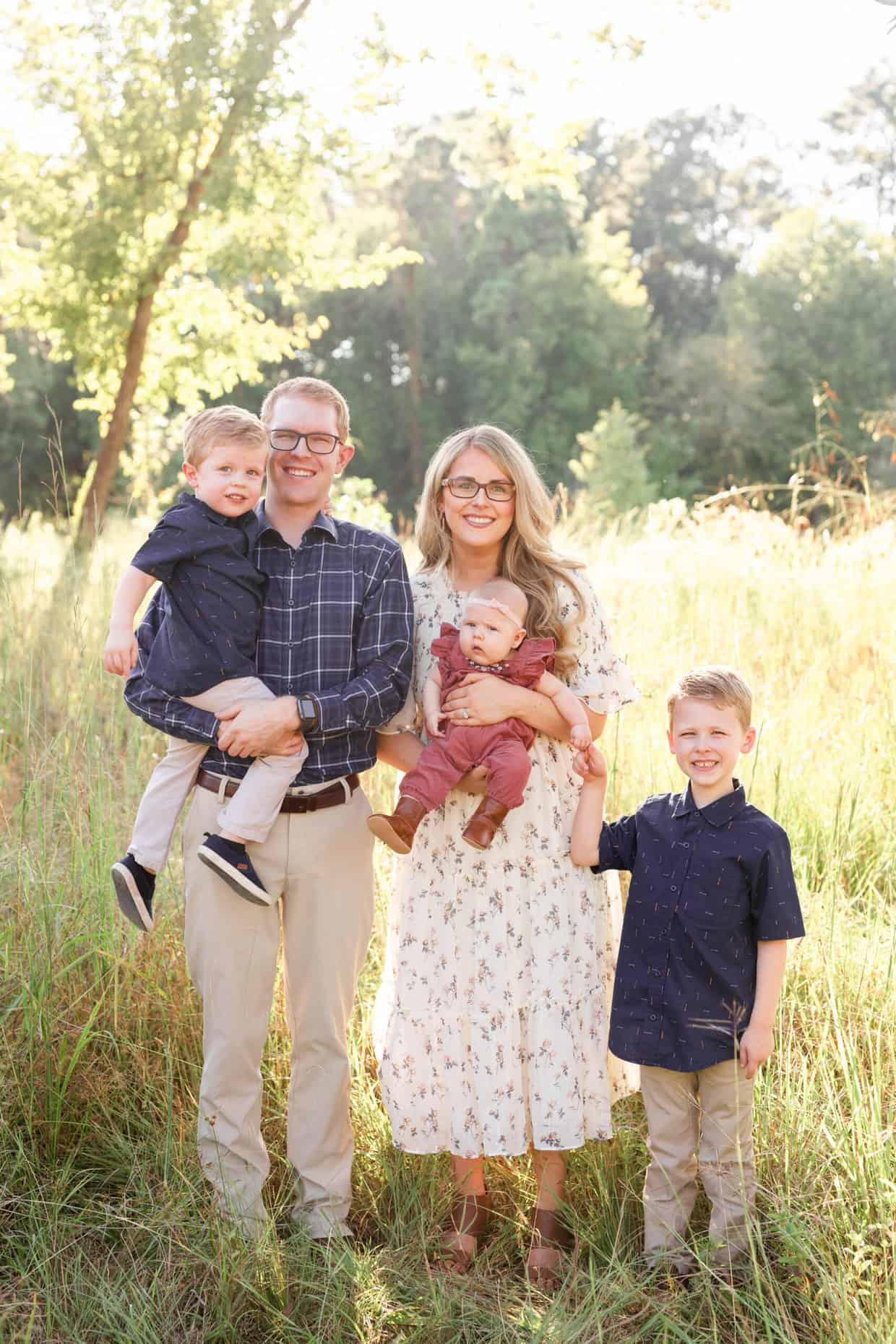 A husband and wife wearing glasses, standing in a field with wife holding a baby girl in pink jumper, and two boys in slacks and blue shirts.