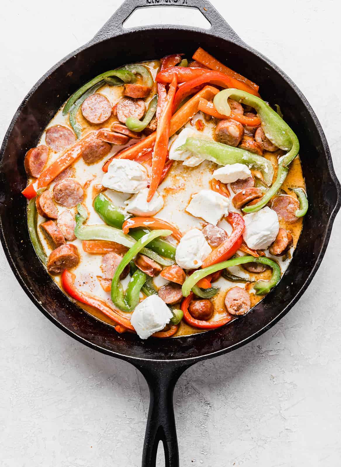 A black skillet with sliced green and red peppers, sausages, and cubed cream cheese, for making Cajun Sausage Pasta.