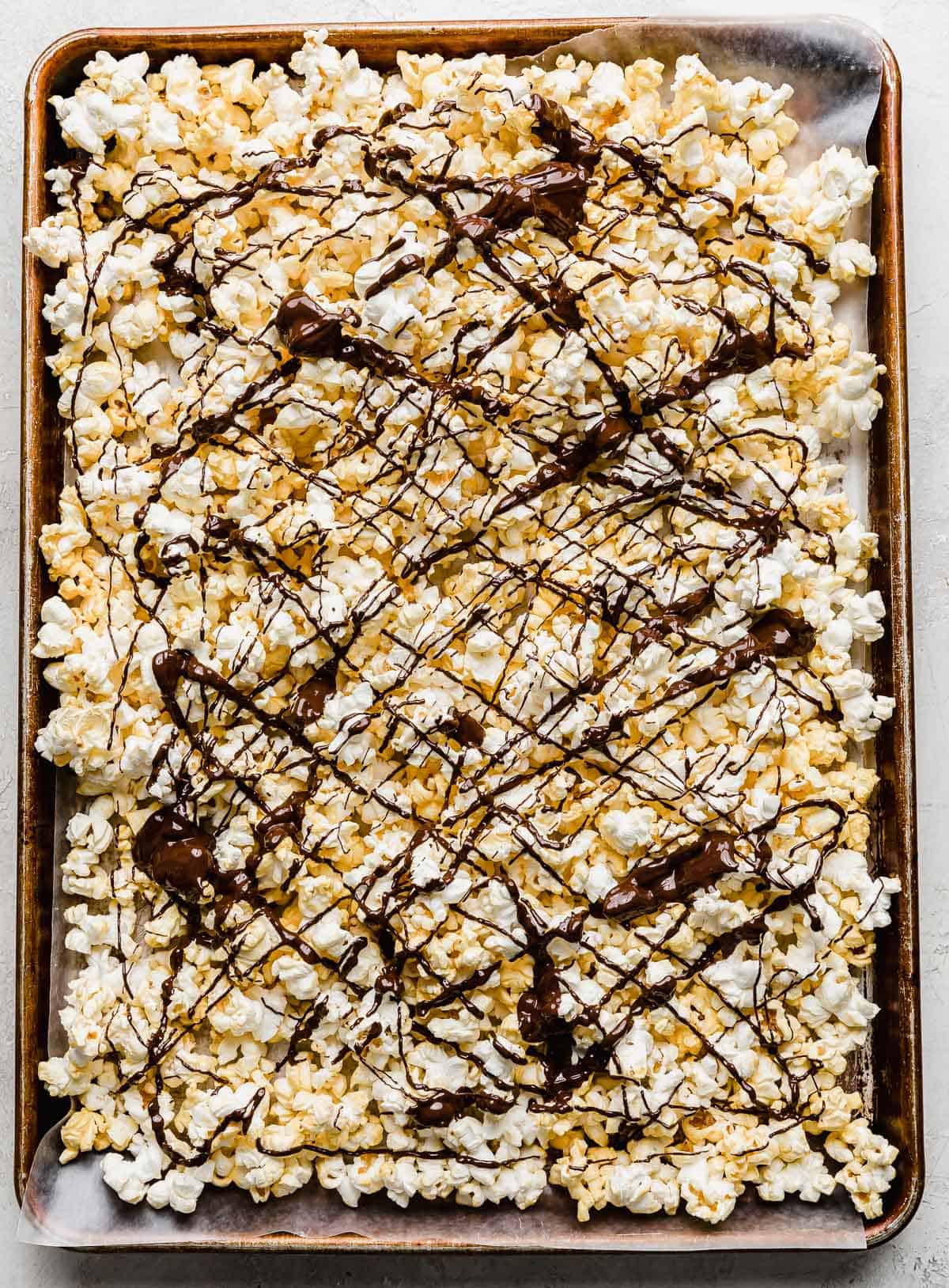 Popped popcorn on a baking sheet with melted dark chocolate drizzled overtop.