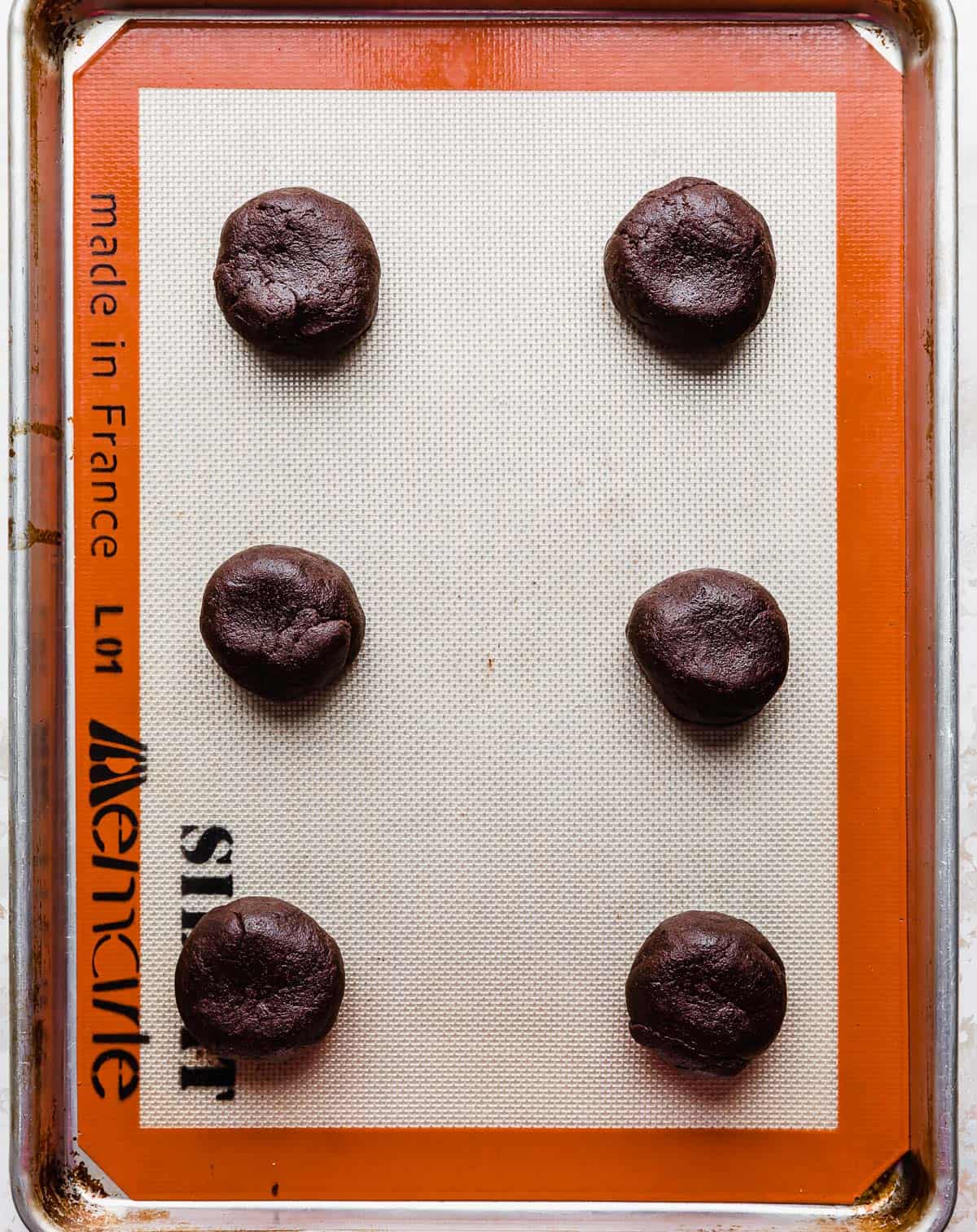 A baking sheet with six chocolate dough balls on it.