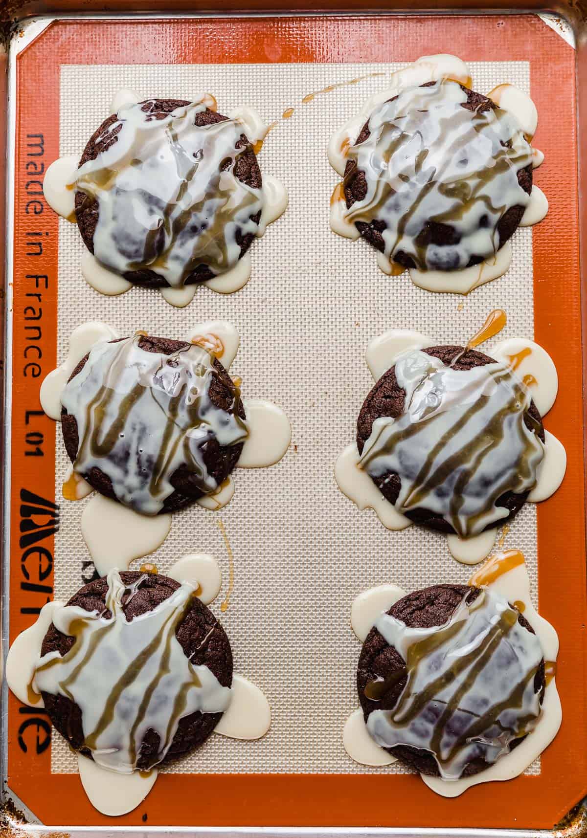 Six chocolate Cookies on a baking sheet with sweetened condensed milk and caramel drizzled overtop each cookie.