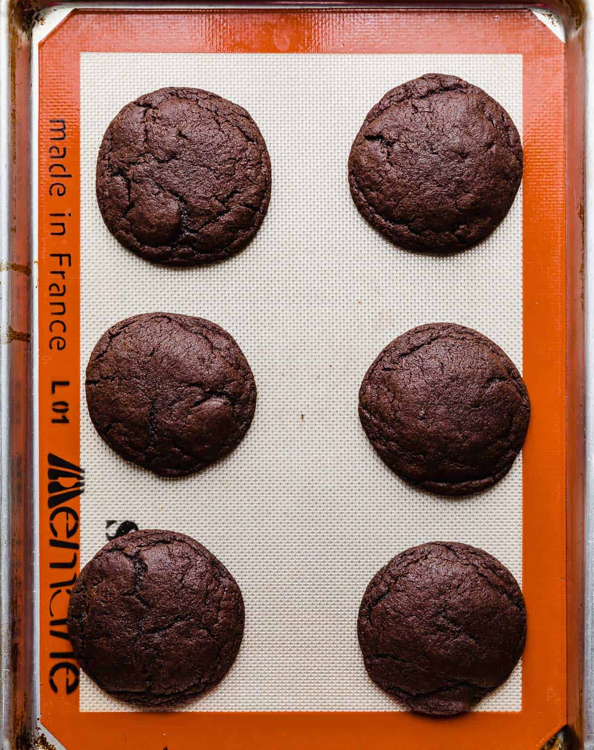 Six baked chocolate cookies on a silicone mat lined baking sheet.