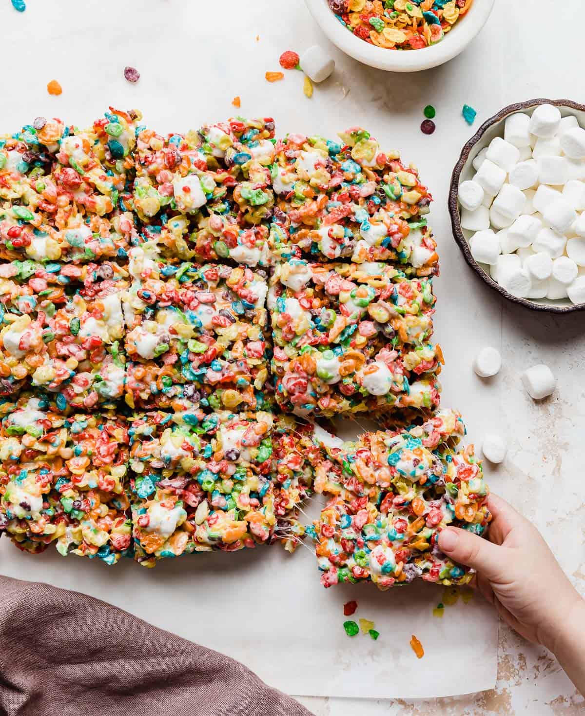 A child's hand grabbing a Fruity Pebble Rice Crispy treat on a parchment paper with mallows around it.