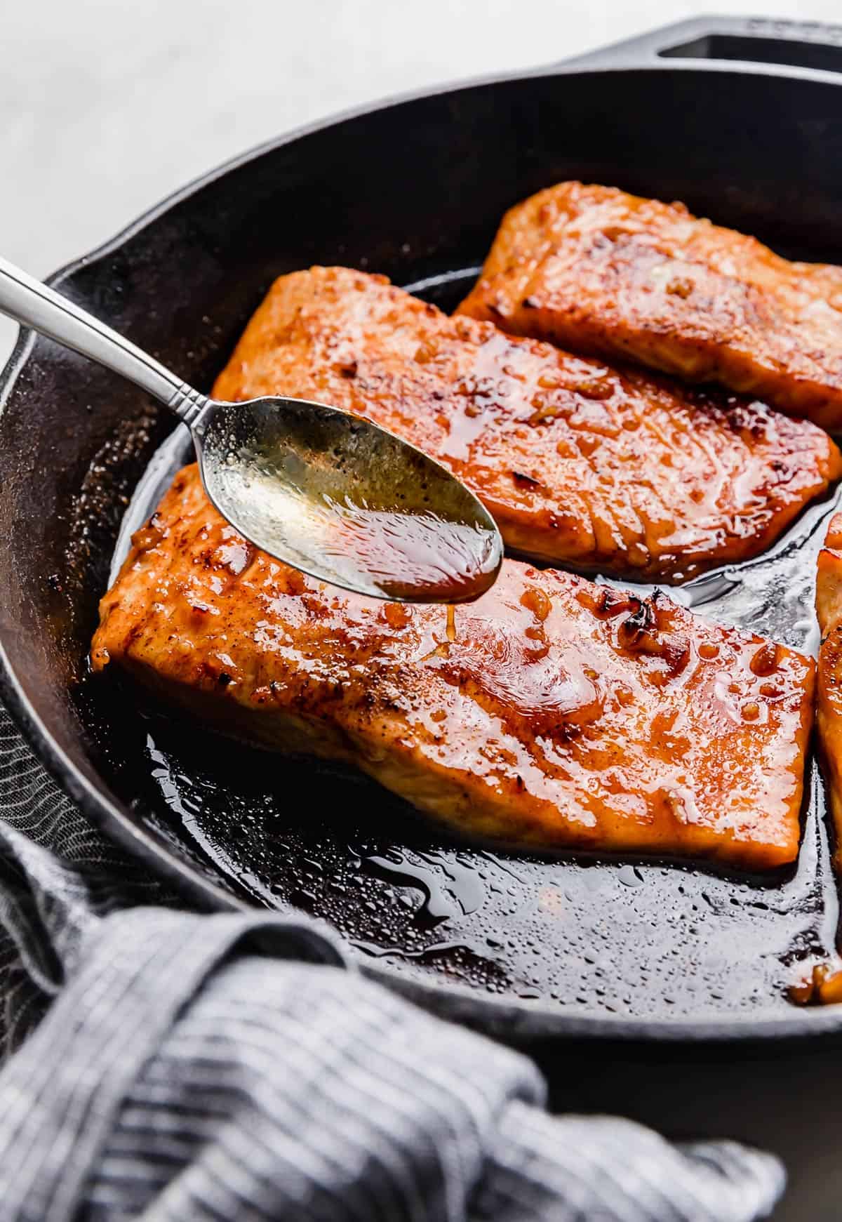 A spoon scooping brown juices over a cast iron Honey Garlic Baked Salmon filet.