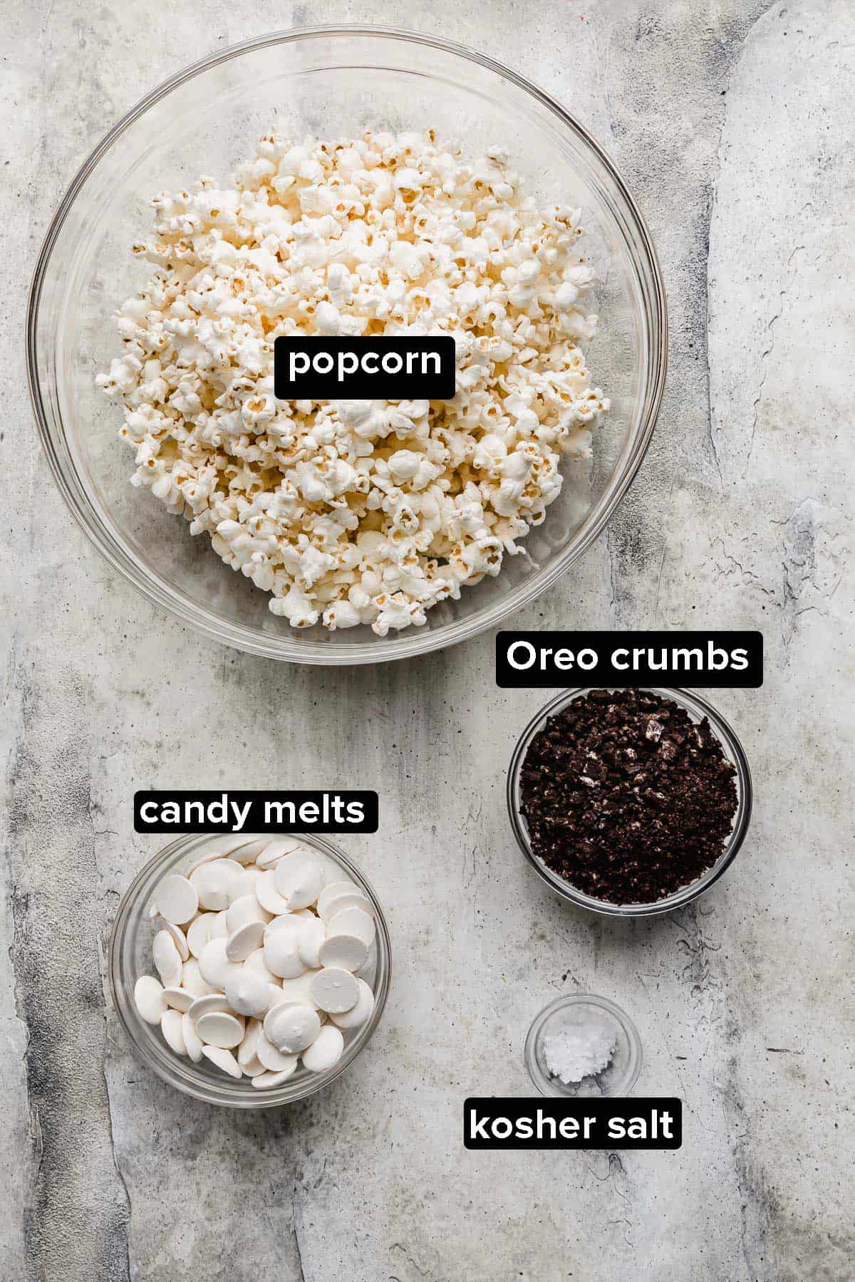 Oreo Popcorn ingredients on a gray background: popcorn, candy melts, oreo crumbs, and kosher salt.