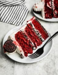 A slice of Oreo Red Velvet Cake on a white plate on a white and gray textured background.