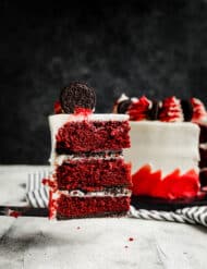 A slice of Oreo Red Velvet Cake being held up in front of the white and red decorated cake.