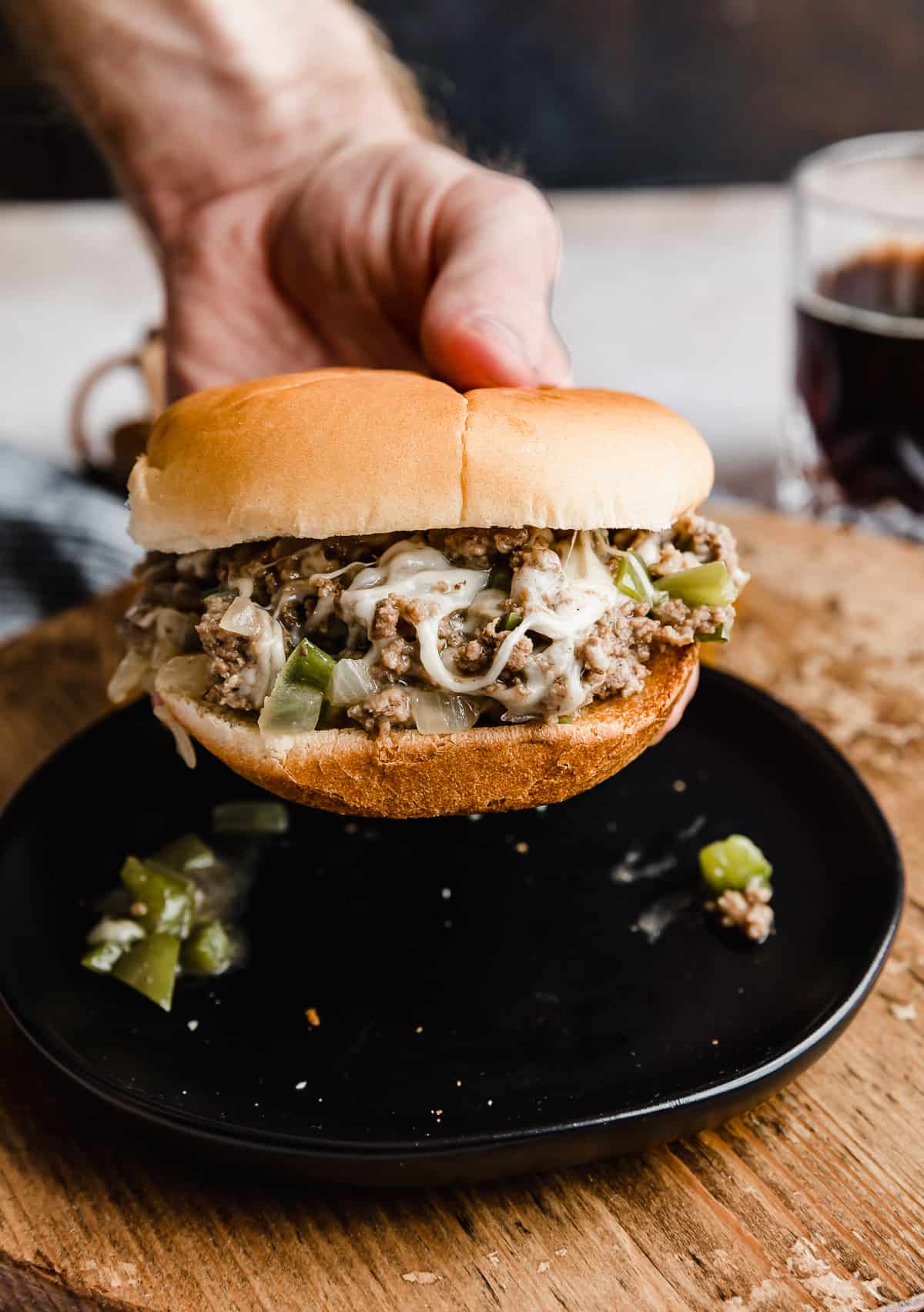 A hand holding a bun topped with Philly Cheesesteak Sloppy Joes hovering over a black plate.