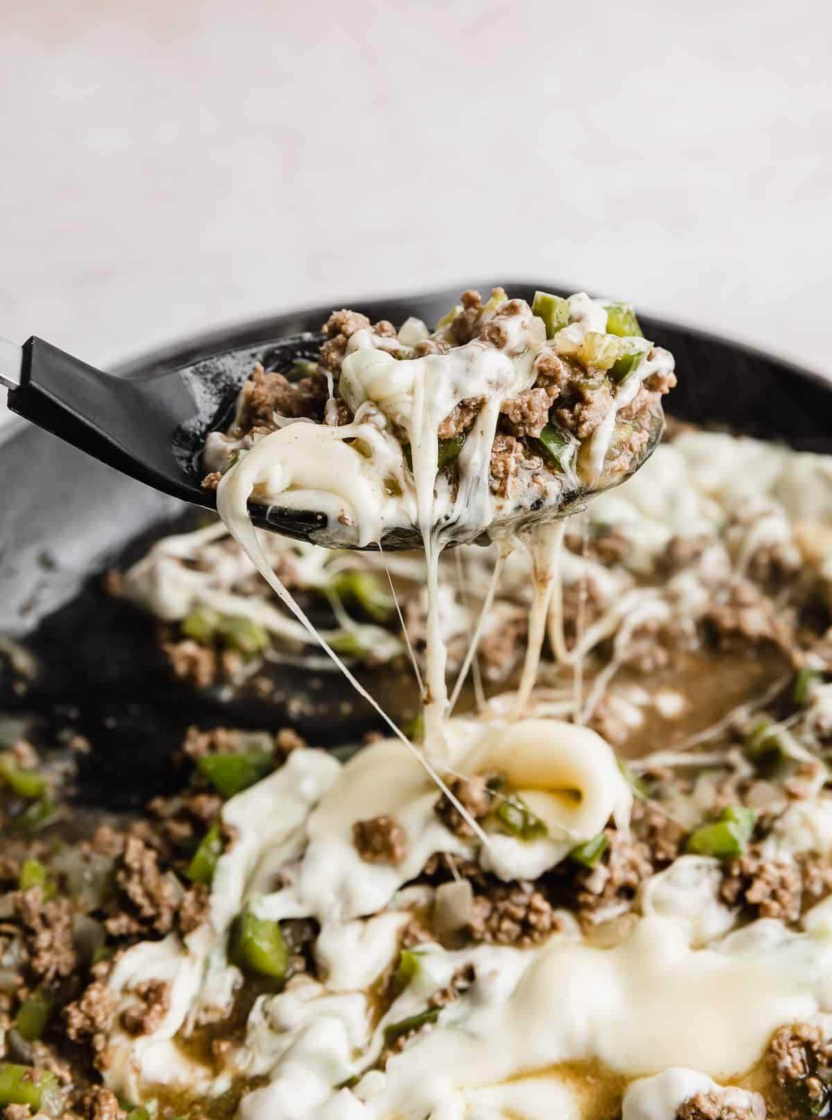 A large black spoon scooping up Philly Cheesesteak Sloppy Joes mixture from a skillet.