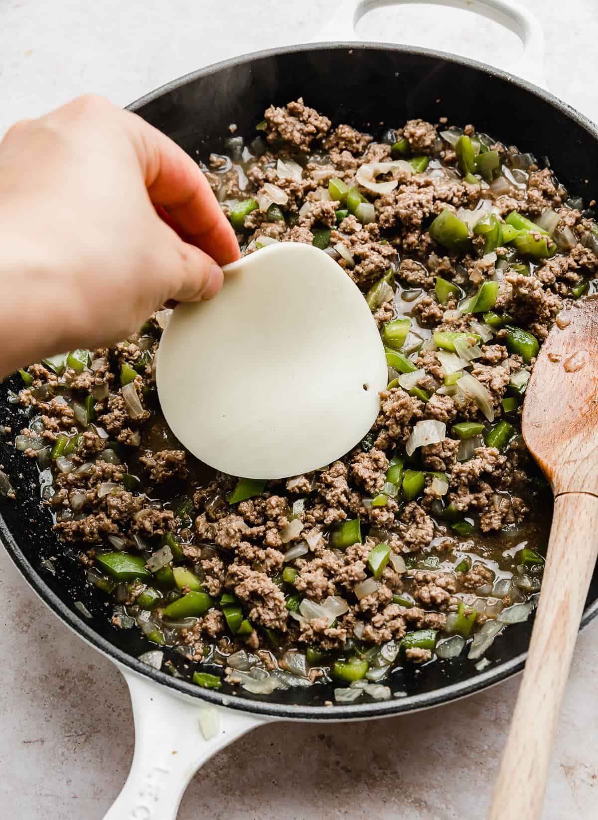 A hand placing a slice of provolone cheese on top of Philly Cheesesteak Sloppy Joes mixture in a skillet.