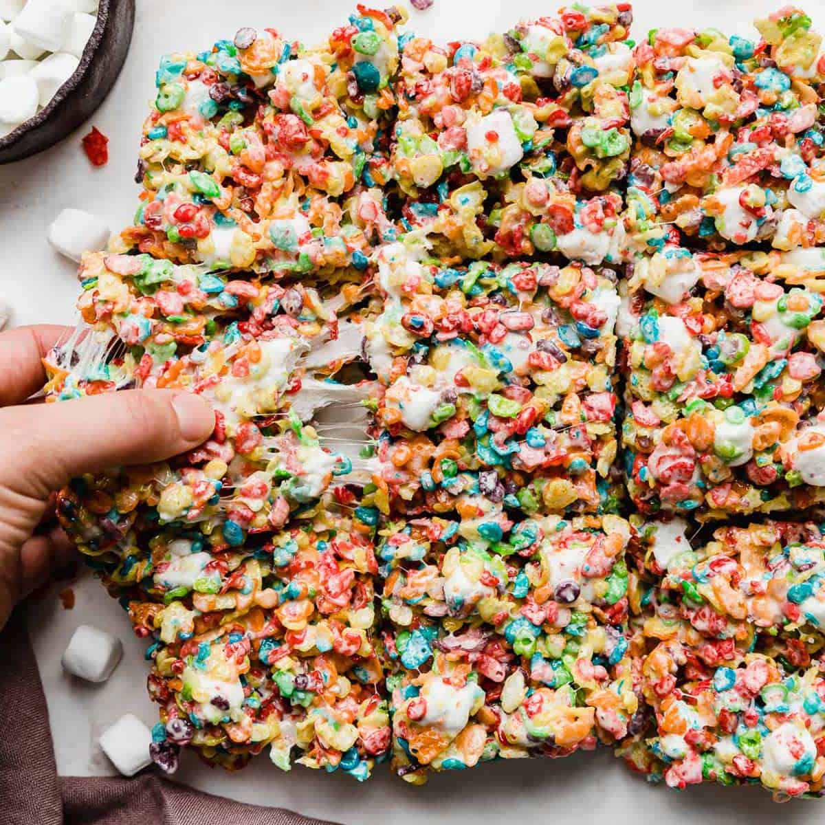 A hand grabbing a cut square of Fruity Pebble Rice Krispies Treats.