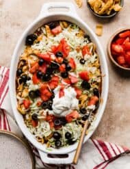 A white oval baking dish filled with Walking Taco Casserole that's topped with chopped tomatoes, olives, and sour cream.