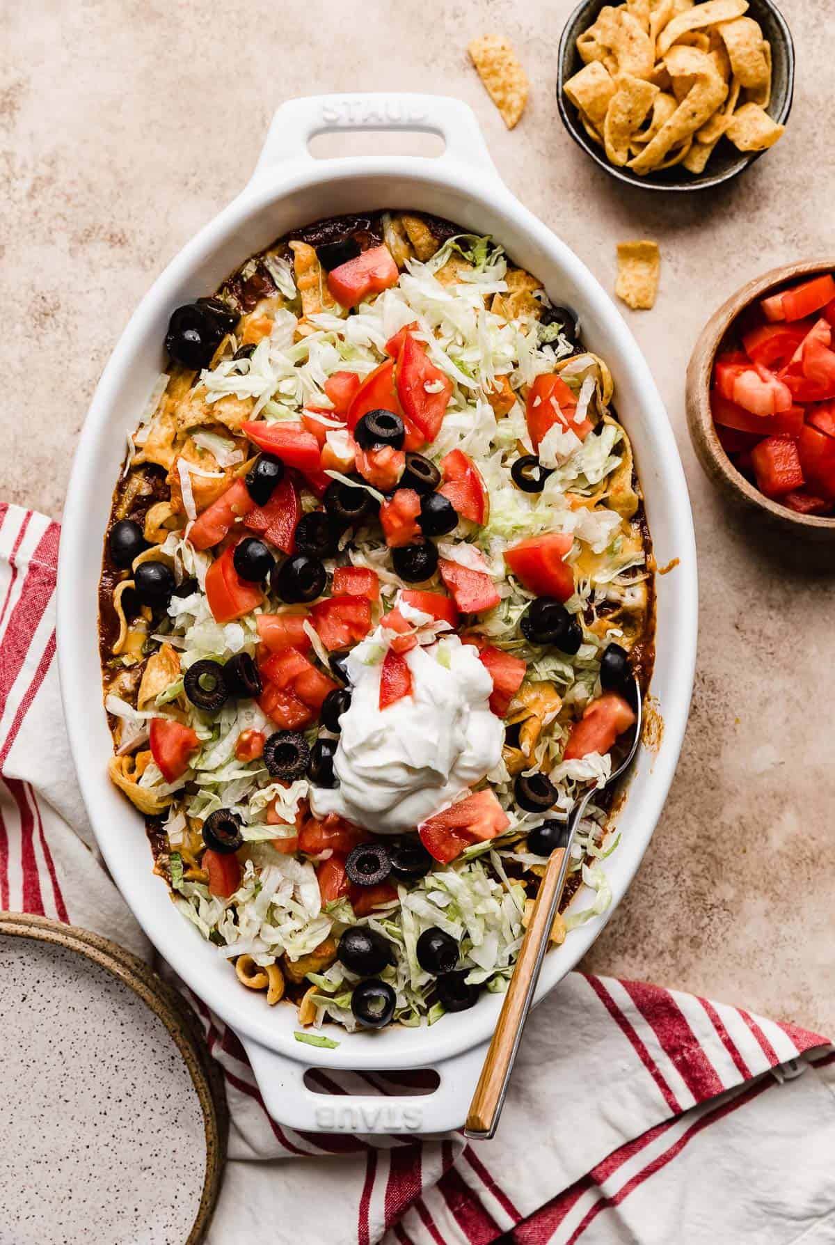 A white oval baking dish filled with Walking Taco Casserole that's topped with chopped tomatoes, olives, and sour cream.