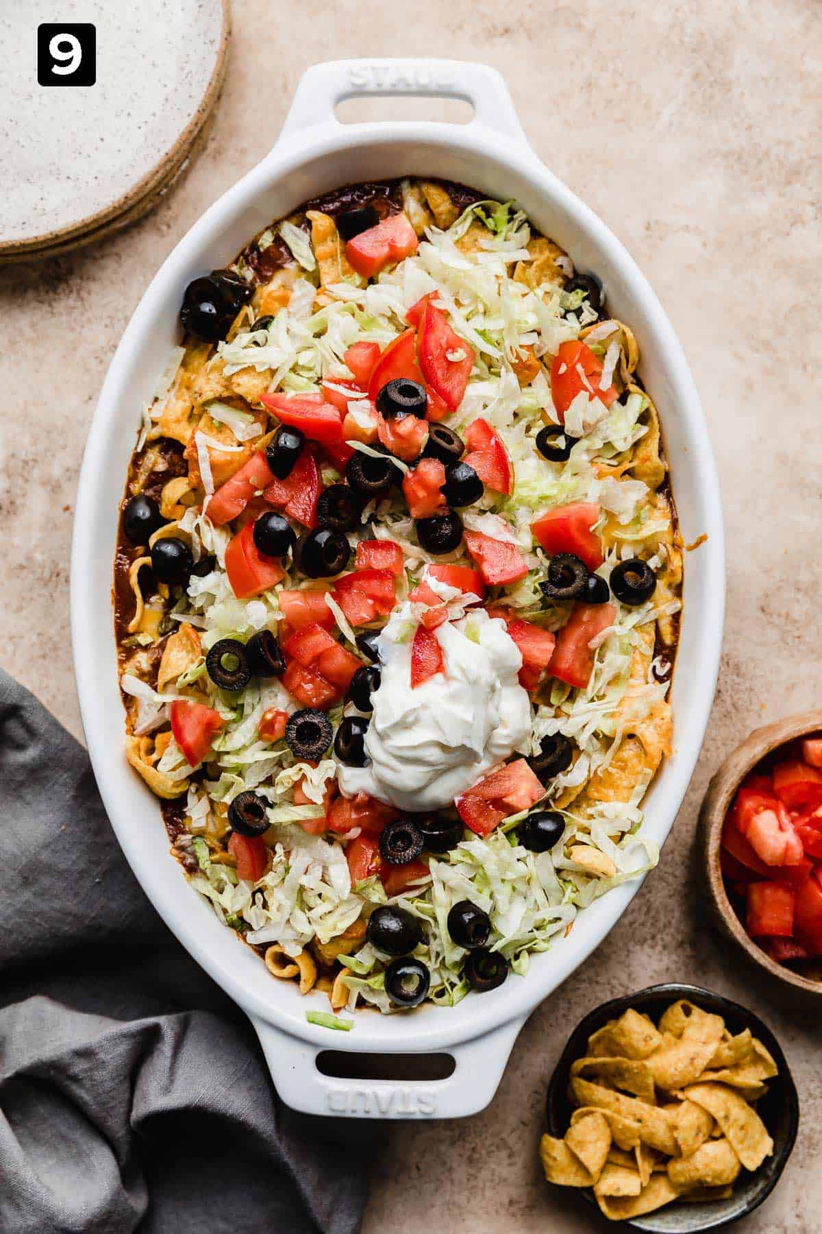 A white oval baking dish filled with a Walking Taco Casserole mixture that's topped with shredded lettuce, tomatoes, olives, and sour cream.