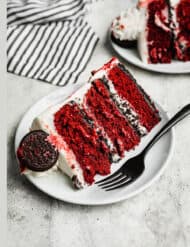 A slice of Oreo Red Velvet Cake with cream cheese frosting on a white plate on a gray background.