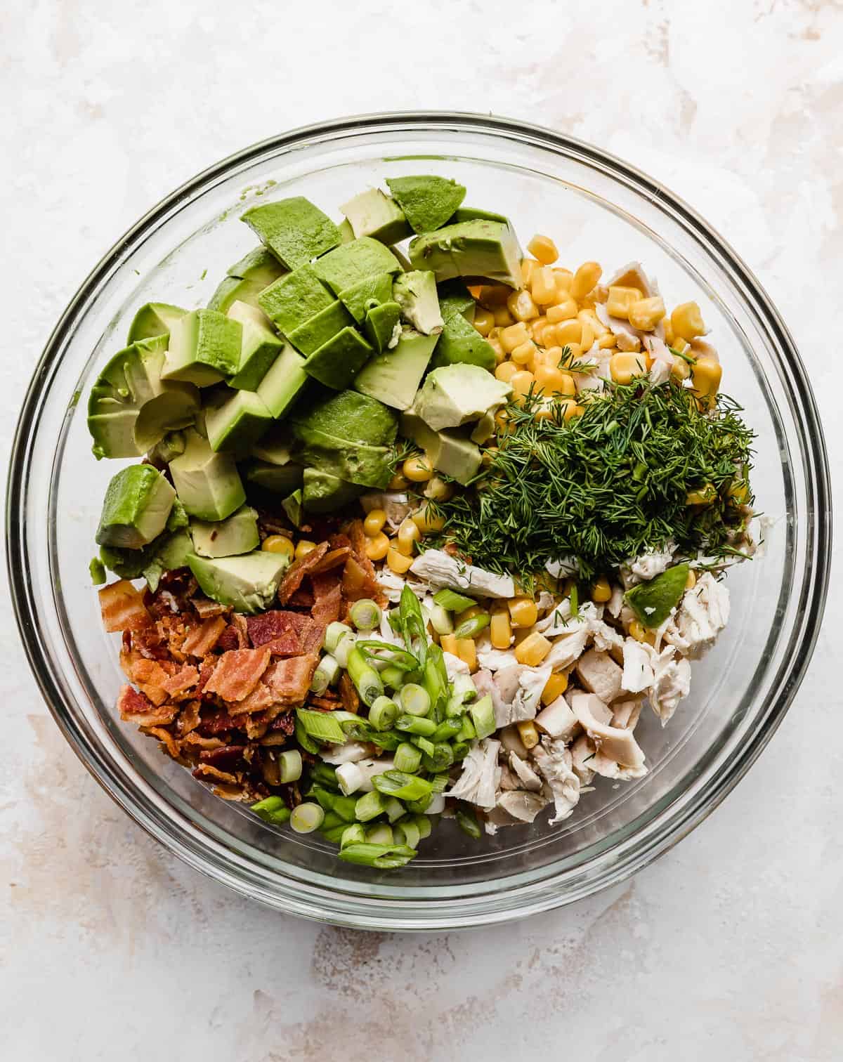 A glass bowl with chopped chicken, avocado, dill, bacon, green onions, and corn in it.
