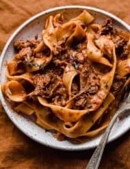A white plate with the best beef Ragu made with pappardelle noodles, on a burnt orange background.