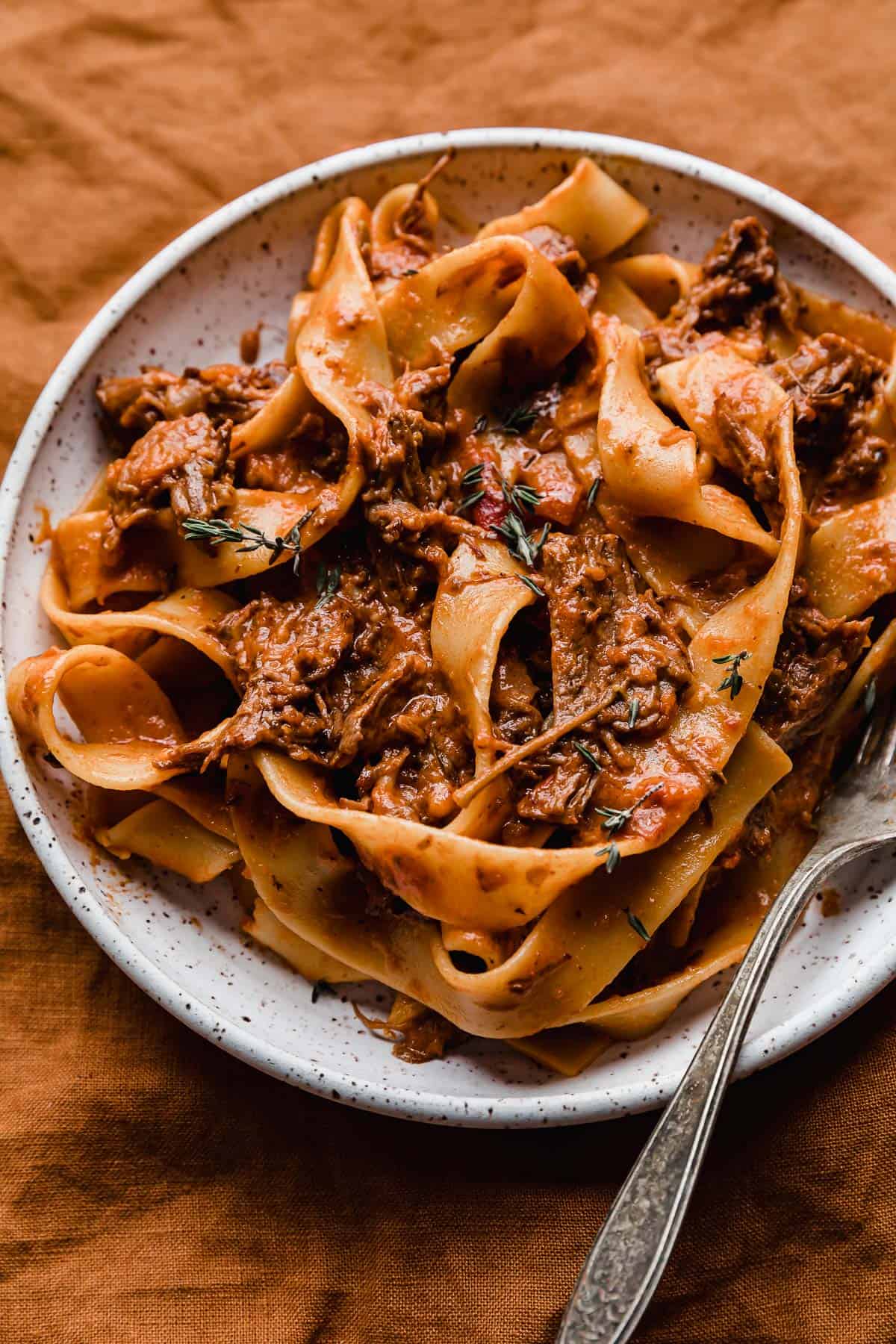 A white plate with the best beef Ragu made with pappardelle noodles, on a burnt orange background.