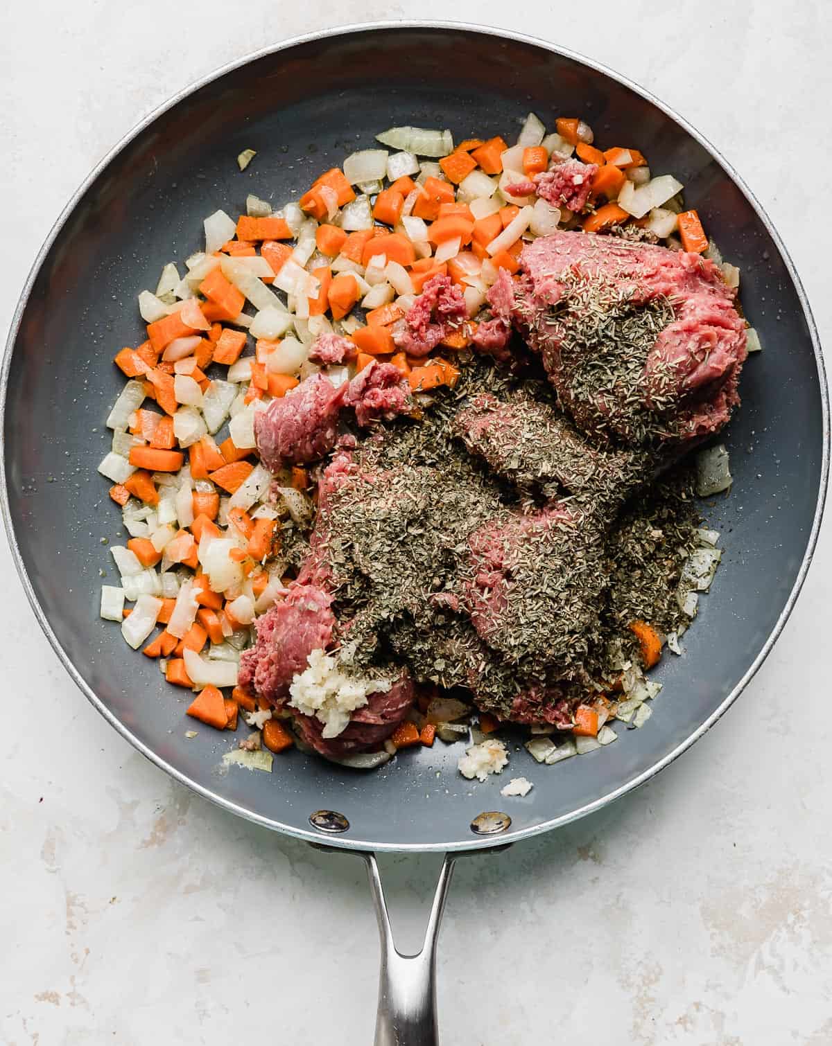 A gray skillet with cooked carrots and onions topped with raw ground beef and seasonings.