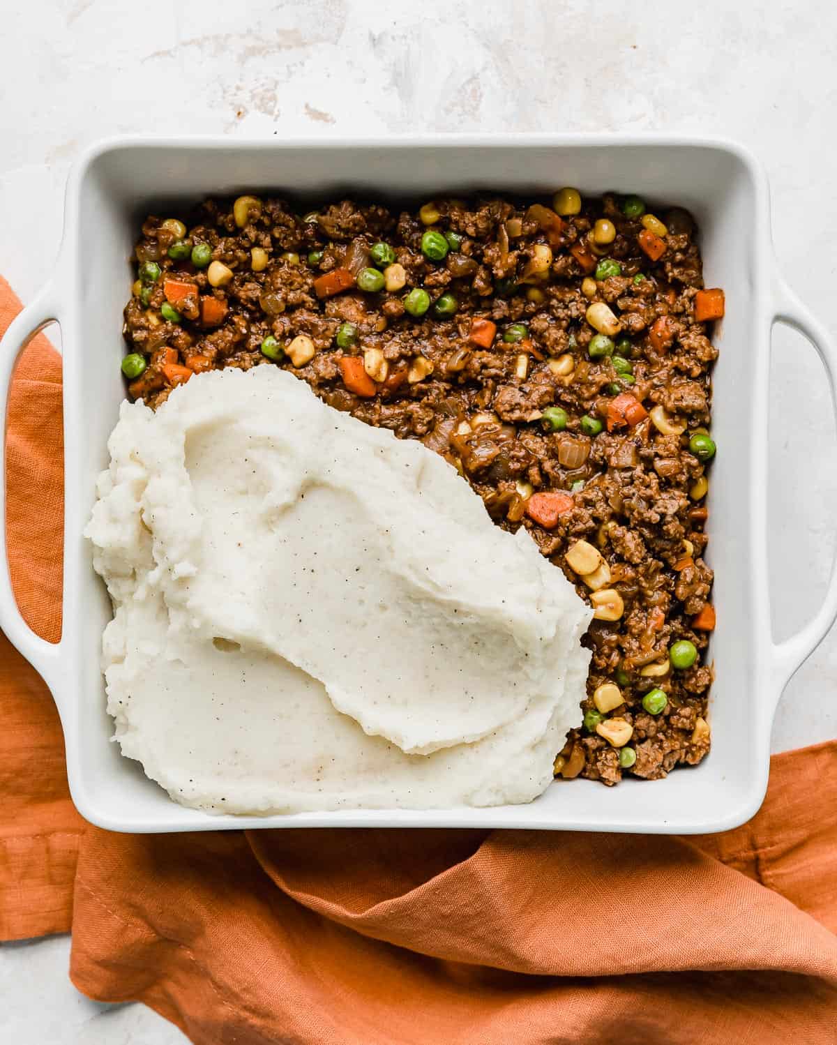 A square baking dish with Easy Shepherd's Pie beef mixture topped with a small portion of mashed potatoes in the bottom left corner of the dish.