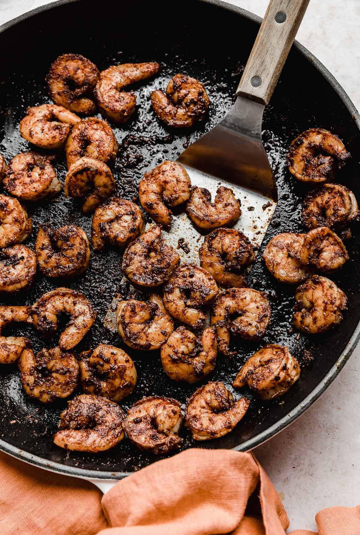 A metal spatula scooping up cooked Blackened Shrimp from a black skillet.
