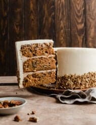 A slice of Carrot Cake with Pineapple and Pecans against a brown background with the cream cheese frosted carrot cake in the background.