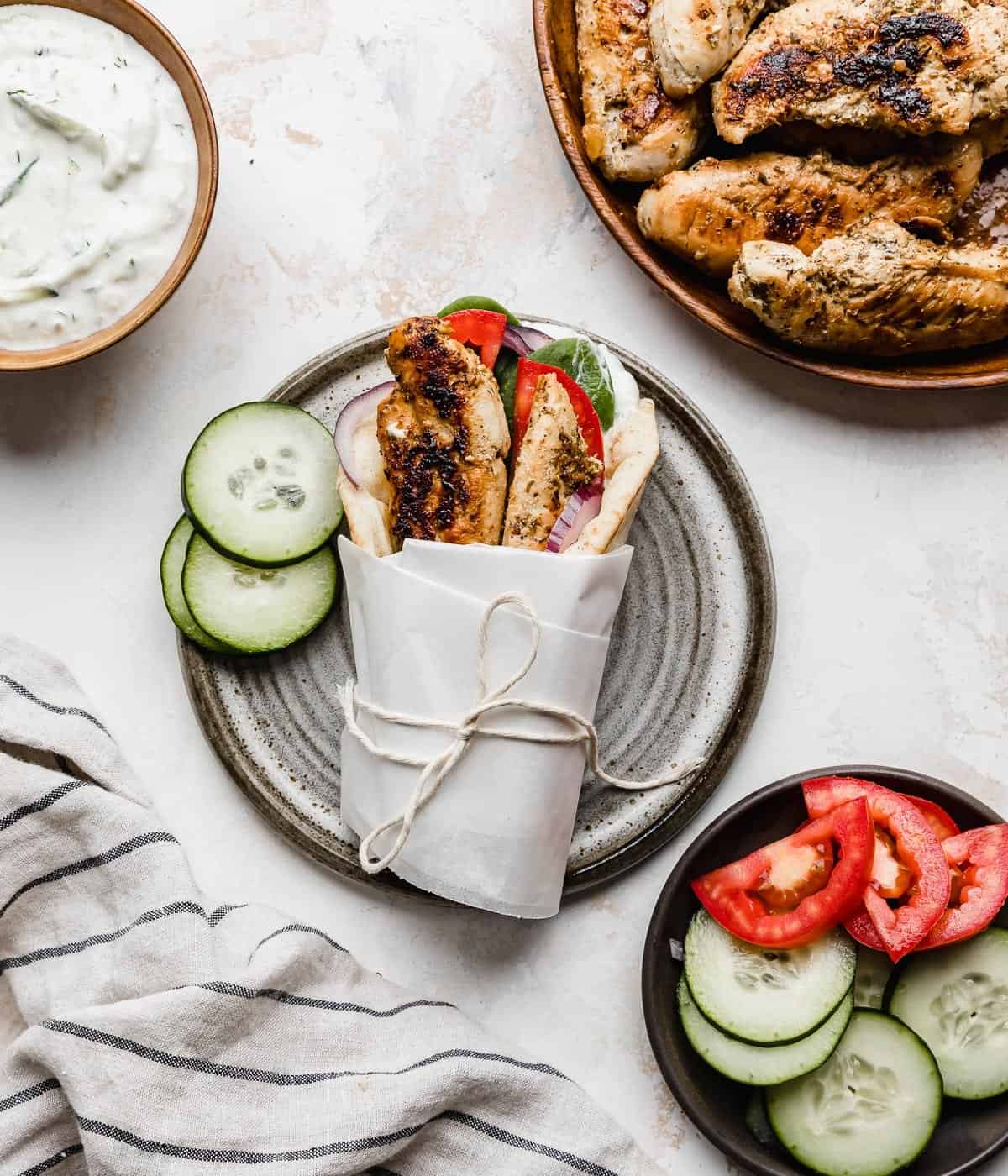 A Greek Chicken Gyro topped with tomatoes, chicken tenders, and wrapped in white parchment on a white background.
