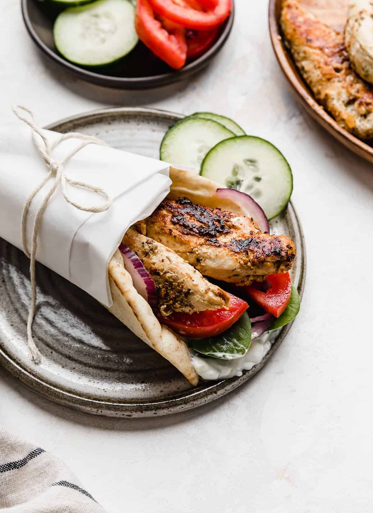 A chicken gyro made with chicken tenders wrapped in white parchment paper, on a grey plate.