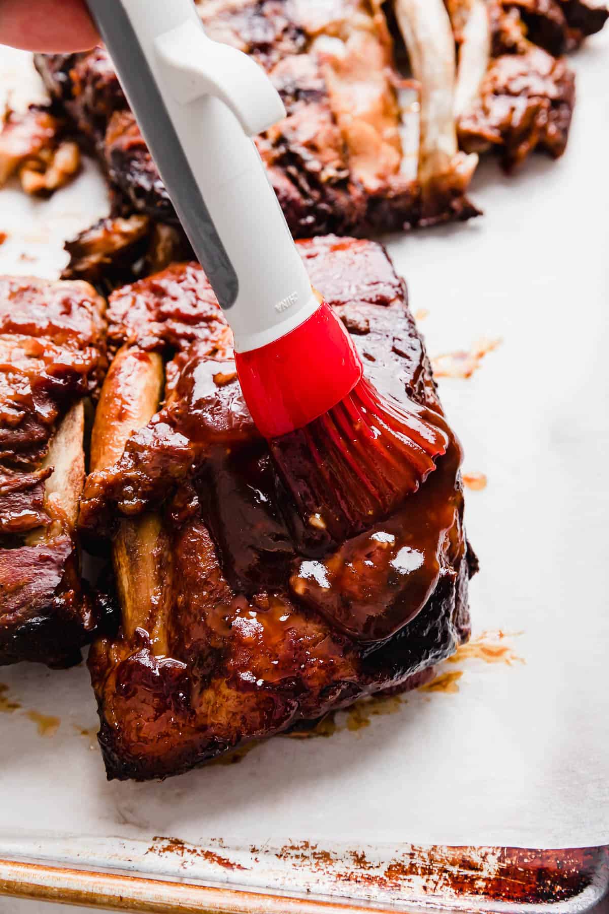 A red pastry brush spreading a dark rib sauce made with Coca Cola, overtop cooked pork ribs.