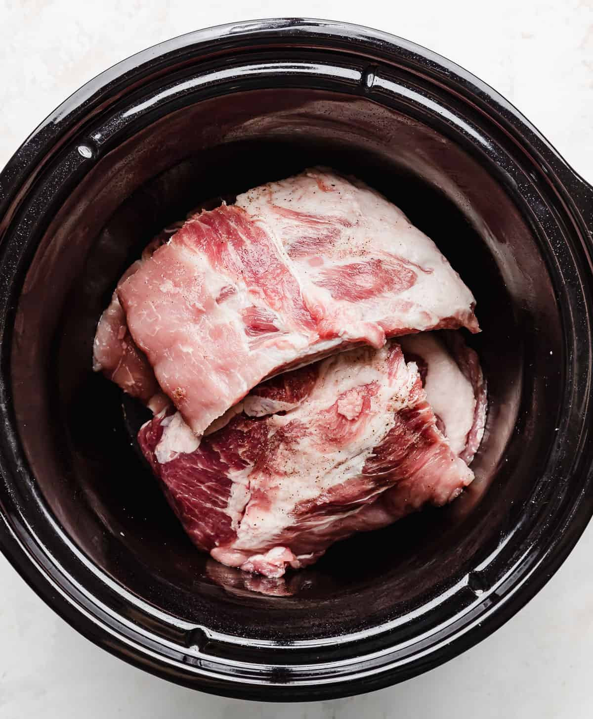 Uncooked baby back pork ribs in a black round slow cooker.