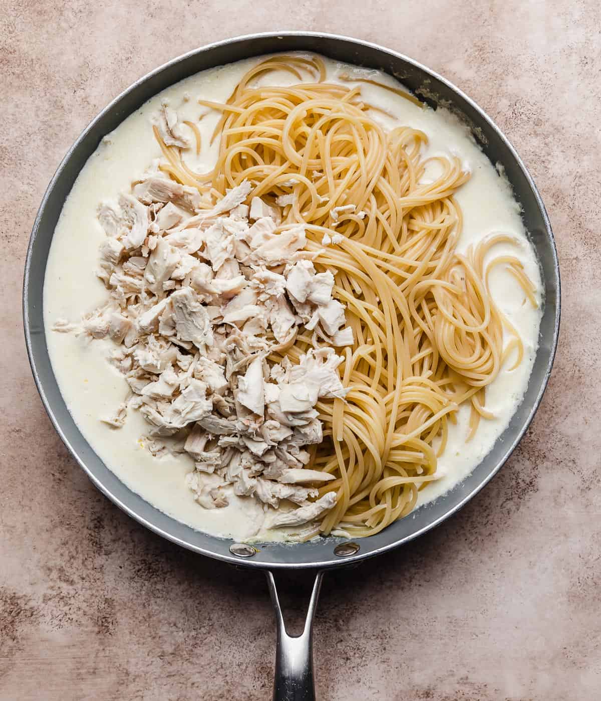 A grey skillet on a beige textured background with a lemon cream sauce, spaghetti noodles and chopped chicken in it.