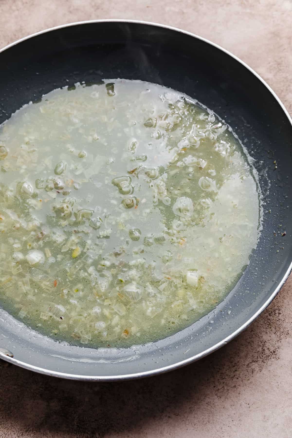 A skillet with chicken broth, diced shallots, and garlic in it.