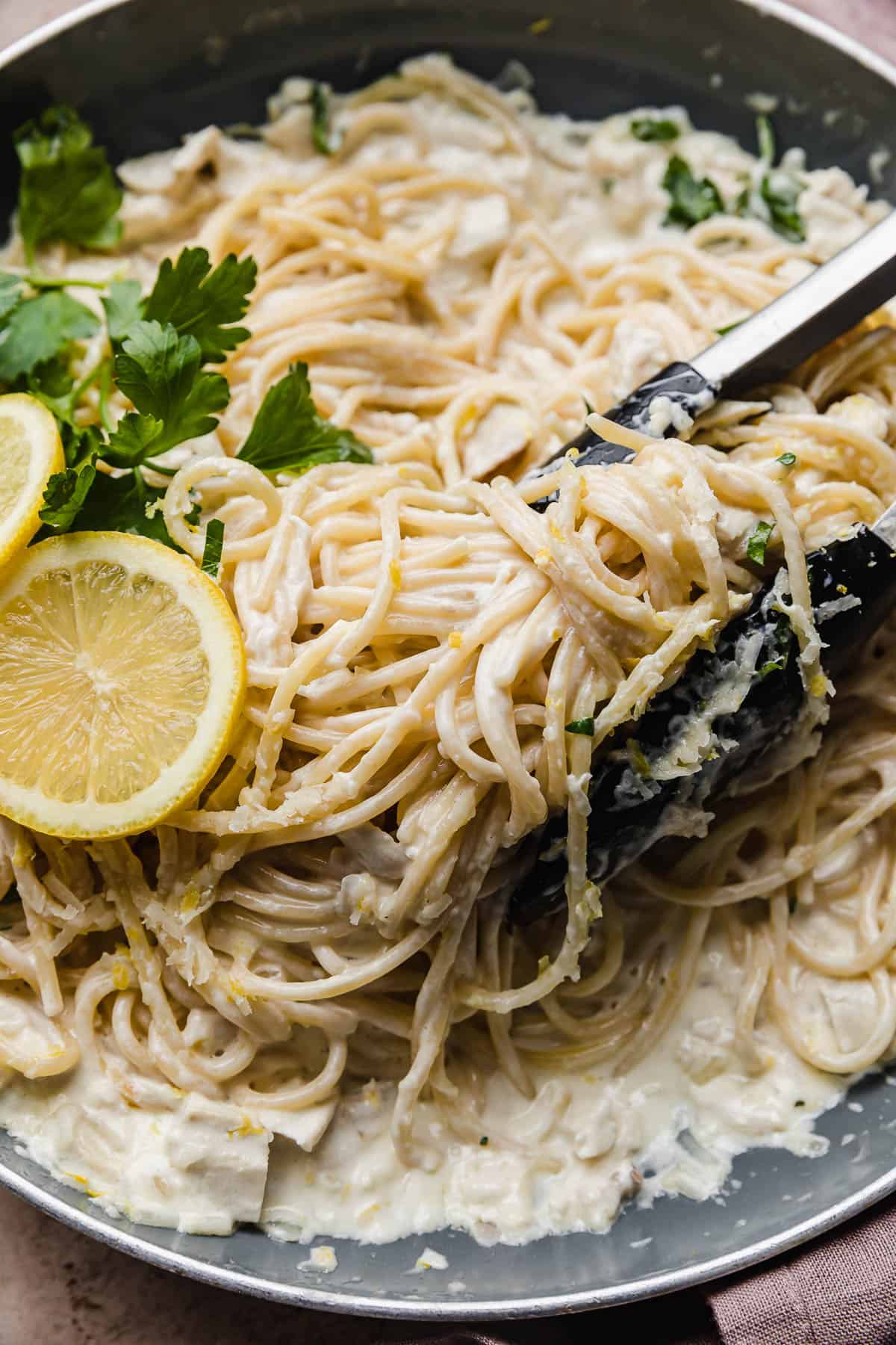 Close up photo of Creamy Lemon Chicken Pasta in a skillet with black tongs pinching some of the spaghetti noodles.