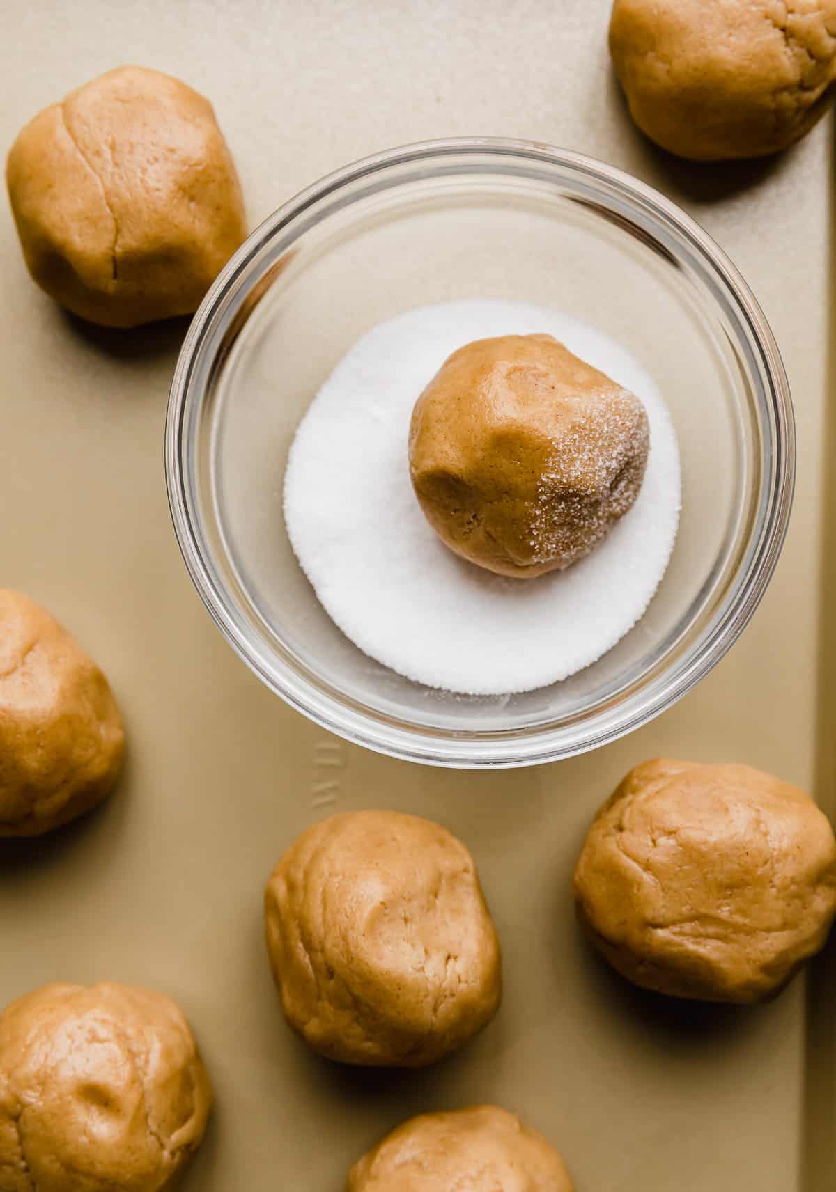 Light brown peanut butter cookies rolled in balls with one ball in a glass bowl that's filled with white sugar.