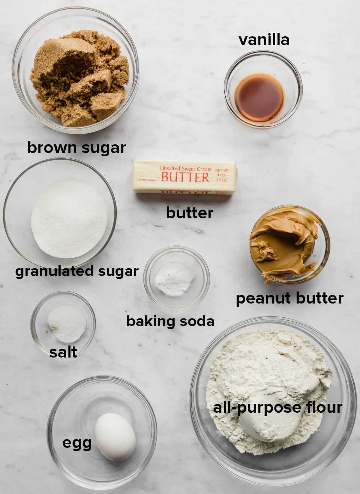 Crumbl Classic Peanut Butter Cookies ingredients placed in glass bowls on a white marble background.