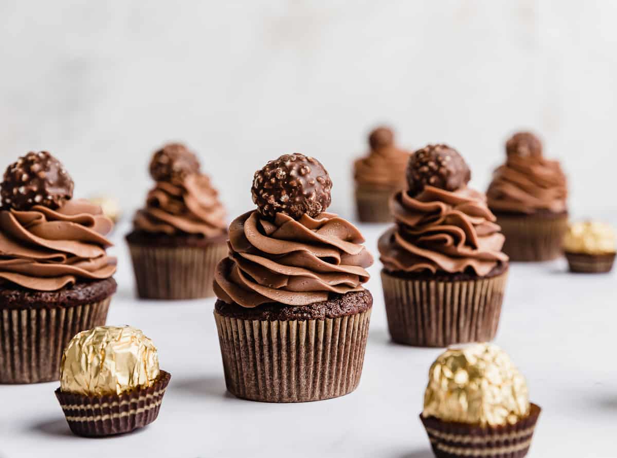 Several Ferrero Rocher Cupcakes topped with a swirl of chocolate frosting and a Ferrero Rocher candy on top, against a white background.