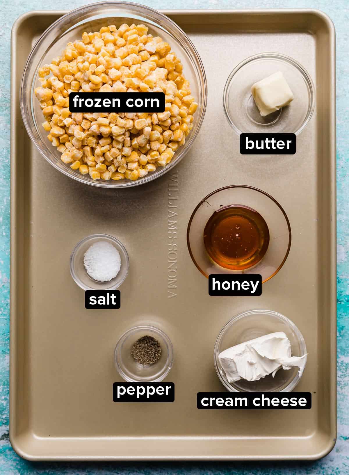 A bronze baking sheet with glass bowls filled with frozen corn, honey, butter, cream cheese, and salt and pepper.