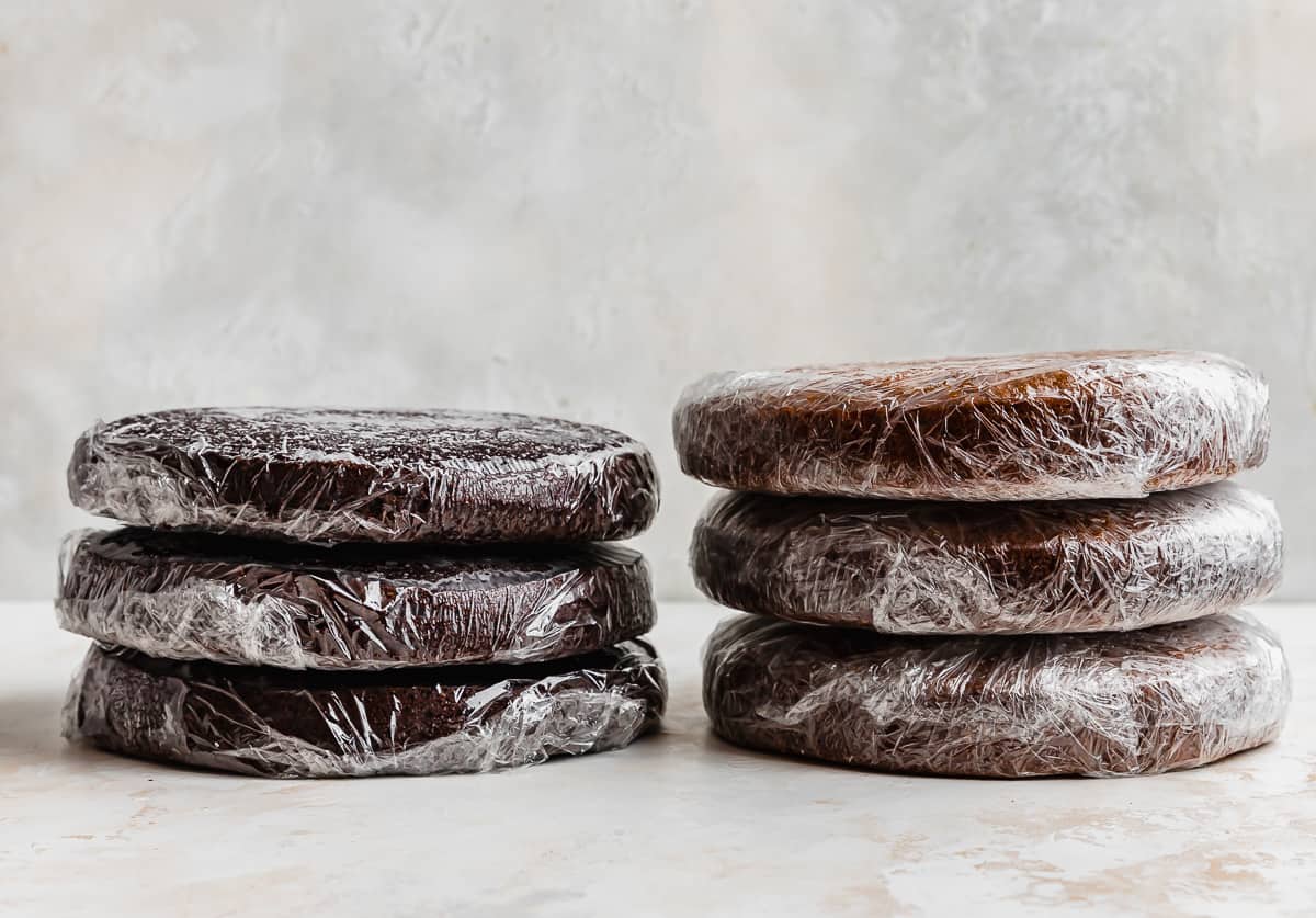 Two stacks of three round cake layers individually wrapped in plastic wrap, against a white textured background.