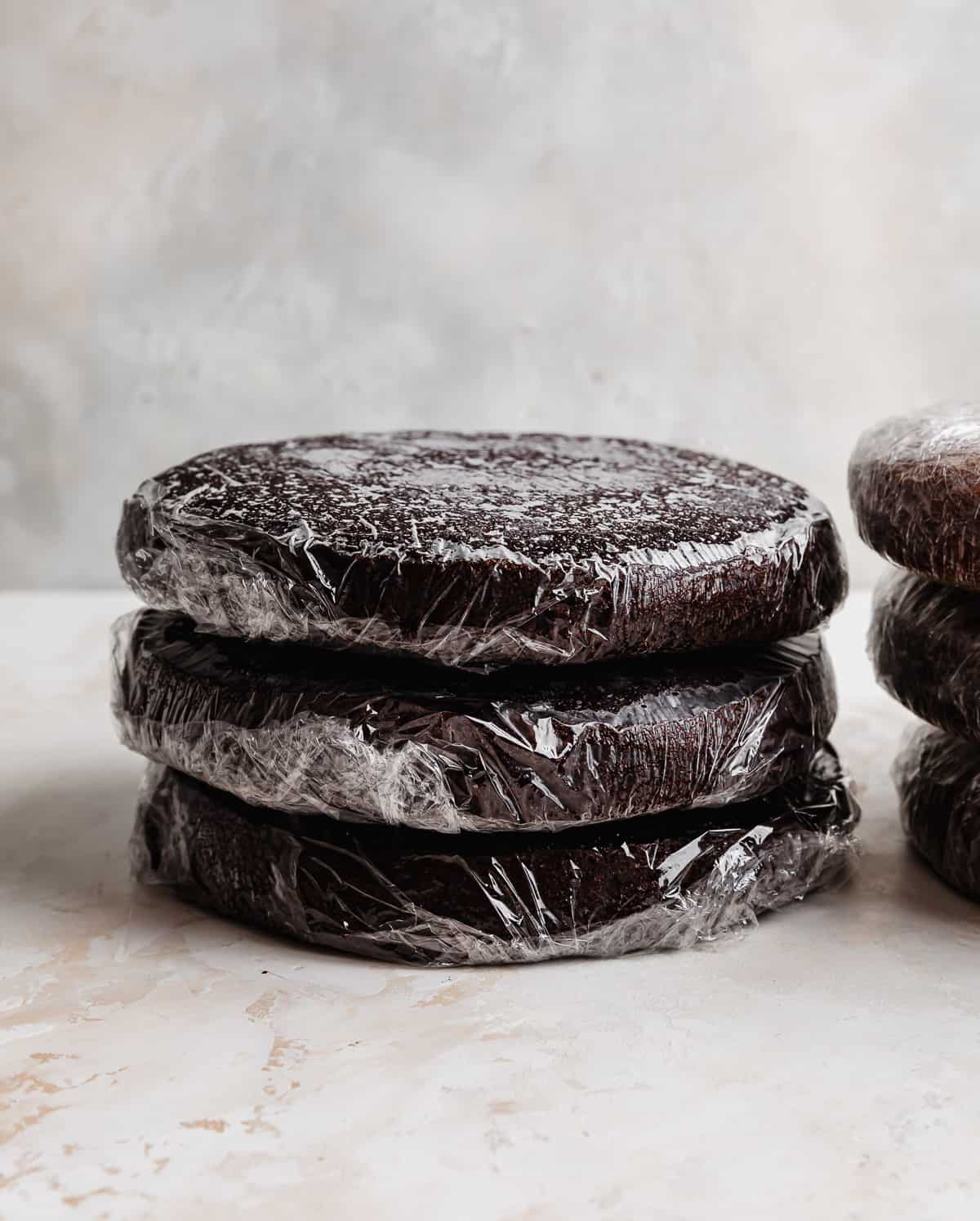Three baked chocolate cake layers individually wrapped in plastic wrap, stacked on top of each other. 