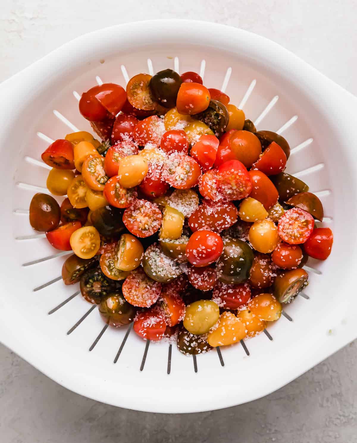 Mixed tomatoes cut into bite size pieces in a white colander with kosher salt sprinkled overtop.