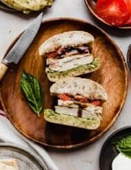 A Chicken Caprese Sandwich Recipe on a brown wooden plate on a beige background with a red and white stripe napkin to the side of the plate.