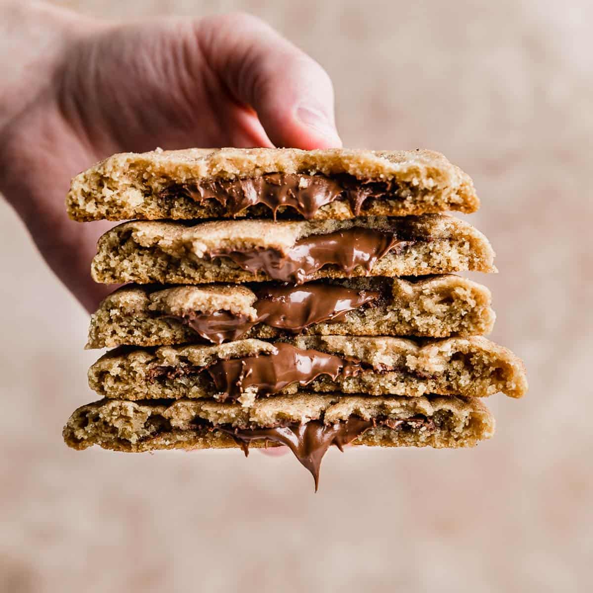 A hand holding a stack of 5 Crumbl Hazelnut Churro Cookies broken in half, against a beige background.