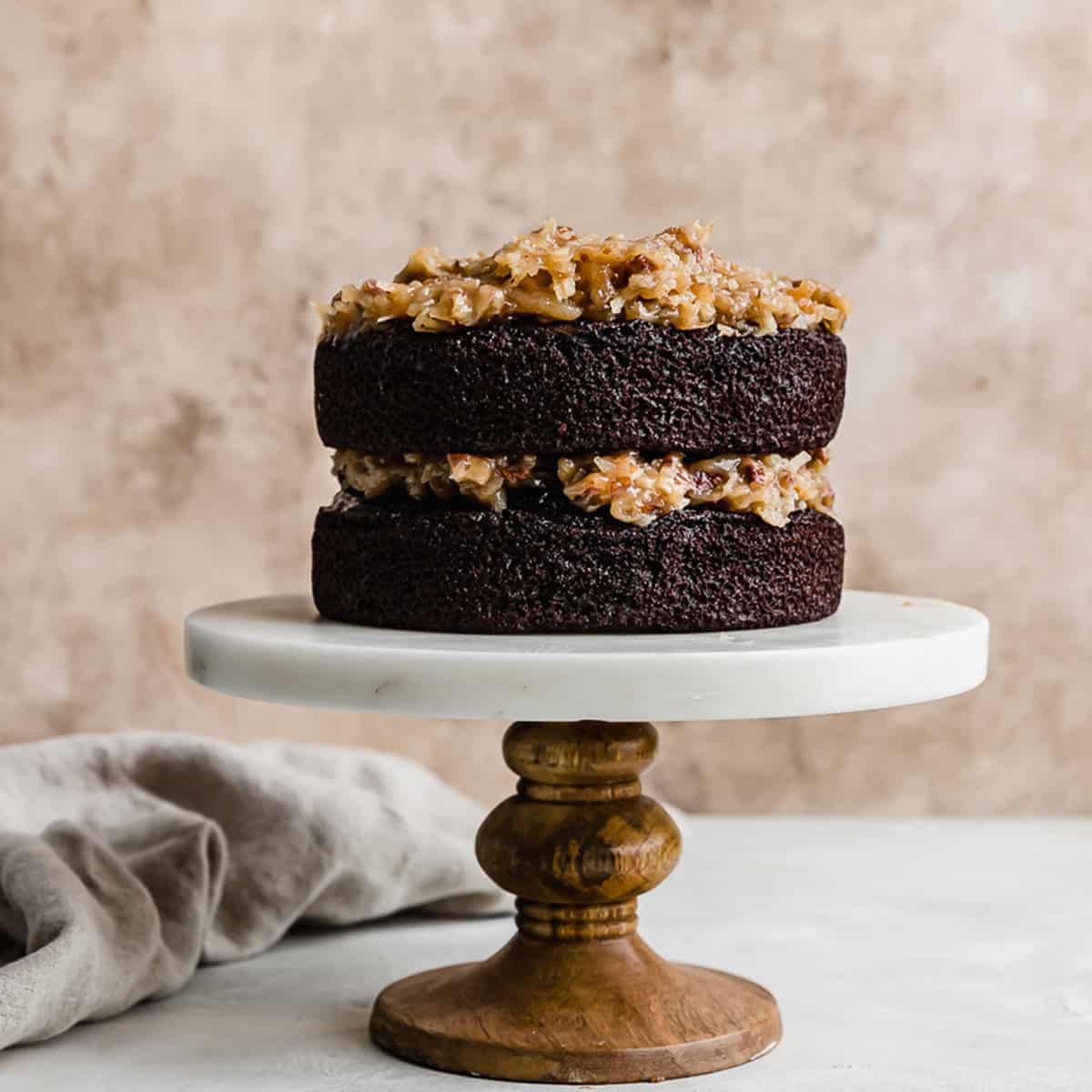 A two layer Small German Chocolate Cake on a white marble cake stand against a light brown background.