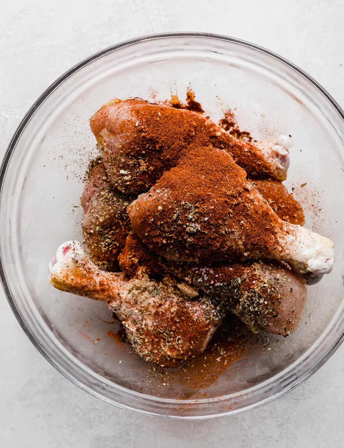 Dried spices such as paprika, Italian seasoning, salt, and pepper sprinkled overtop raw chicken legs that are in a glass bowl.