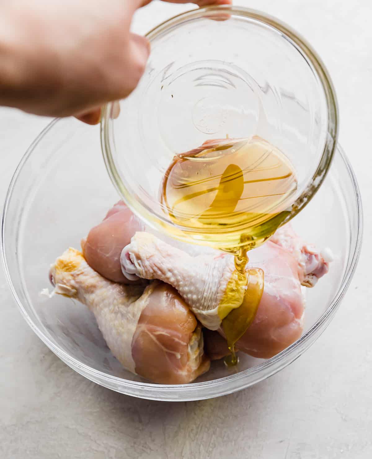 Olive oil being poured overtop chicken drumsticks that are in a glass bowl.
