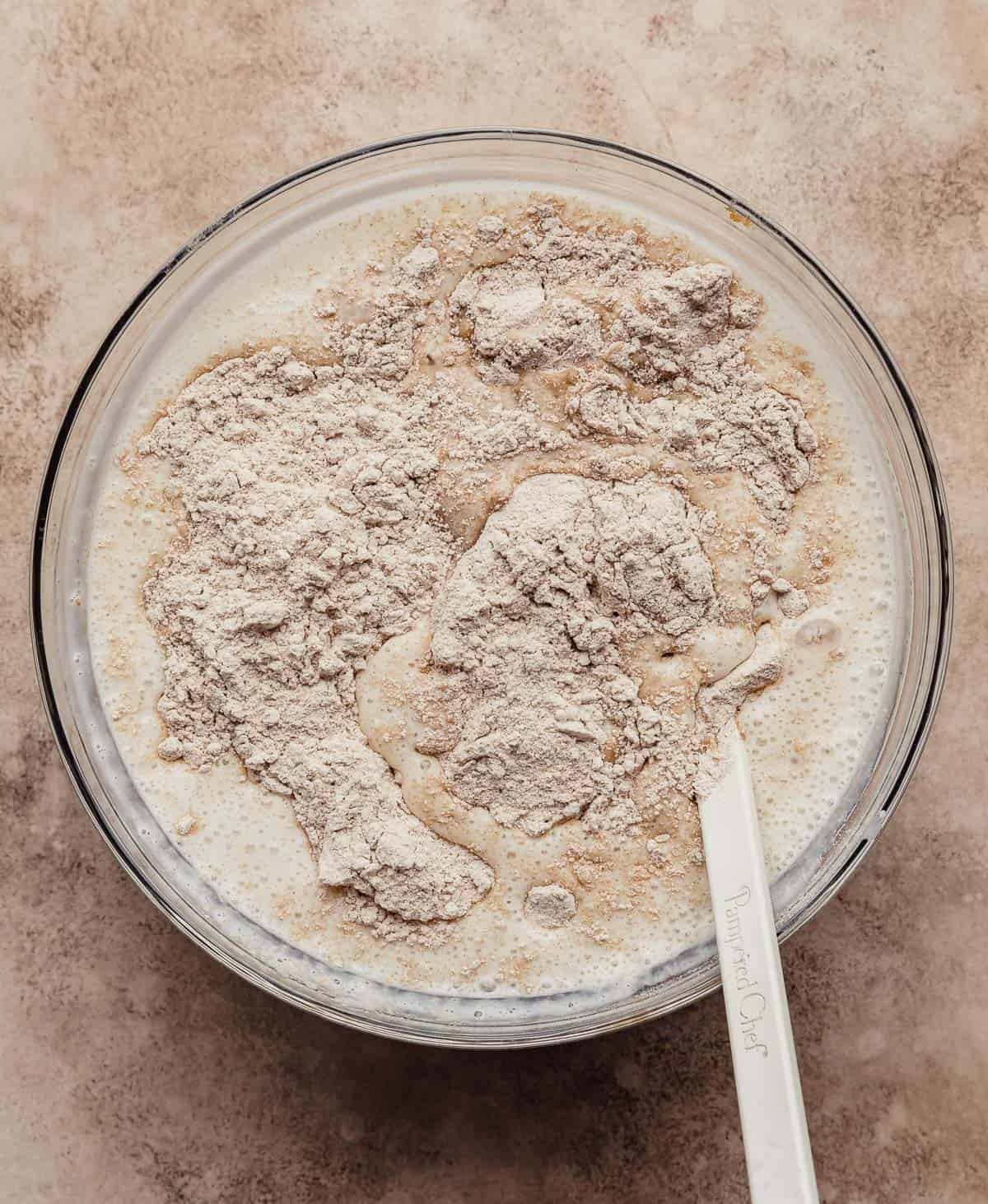 Whole wheat flour in a glass bowl on a light brown textured background.