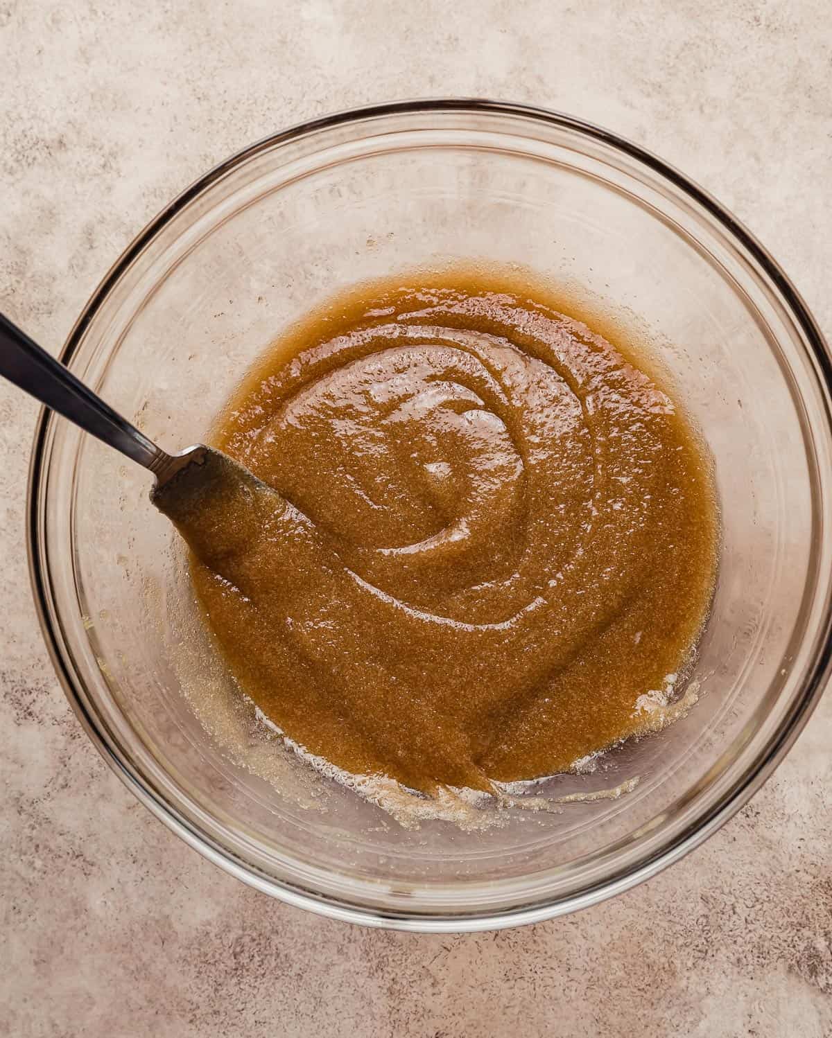 A dark brown colored sugar mixture in a glass bowl on a brown textured background.